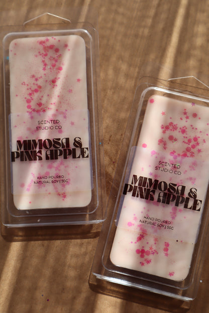 Two scented wax melts in packaging labeled 'Mimosa & Pink Apple' on a wooden surface.