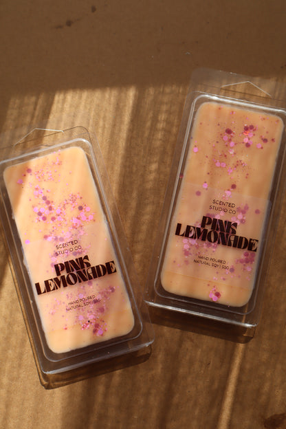 Two pink wax melts with decorative elements in clear packaging on a wooden surface
