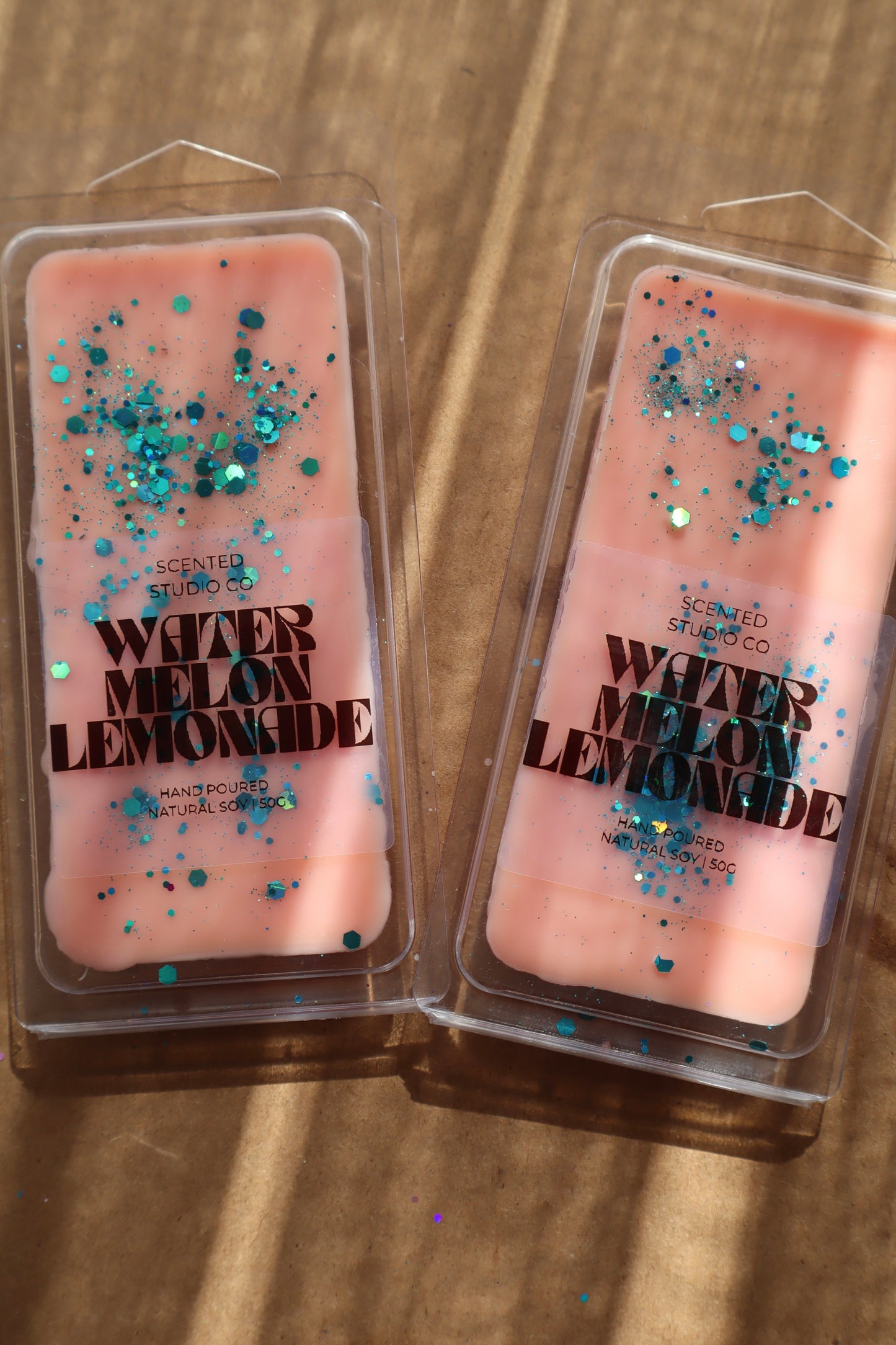 Two pink wax melts with glitter labeled 'Watermelon Lemonade' on a wooden surface.