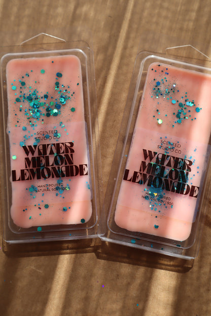 Two pink wax melts with glitter labeled 'Watermelon Lemonade' on a wooden surface.
